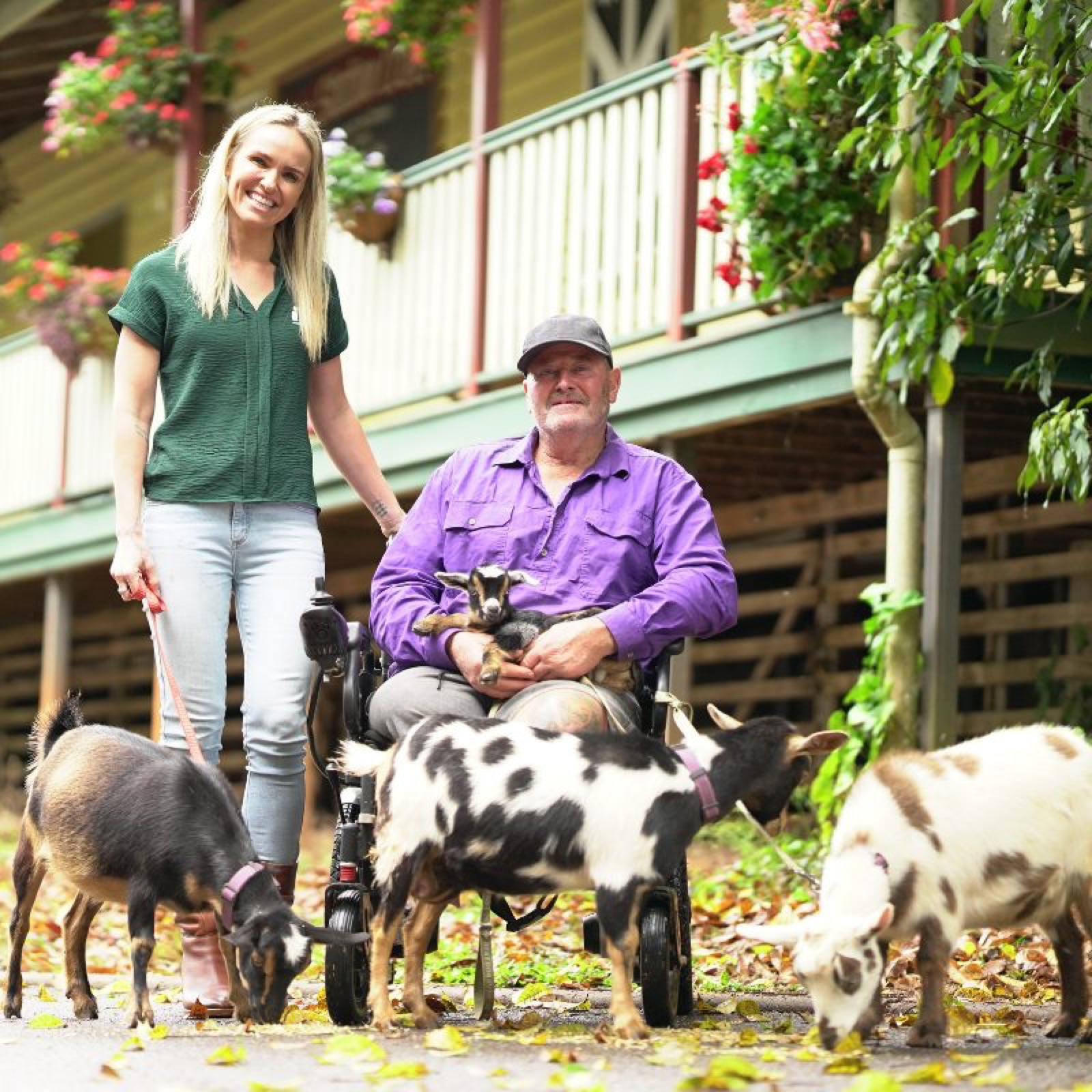 An i'ara participant in his wheelchair cradling a baby goat beside his Support Coordinator, with a classic Queenslander house behind them